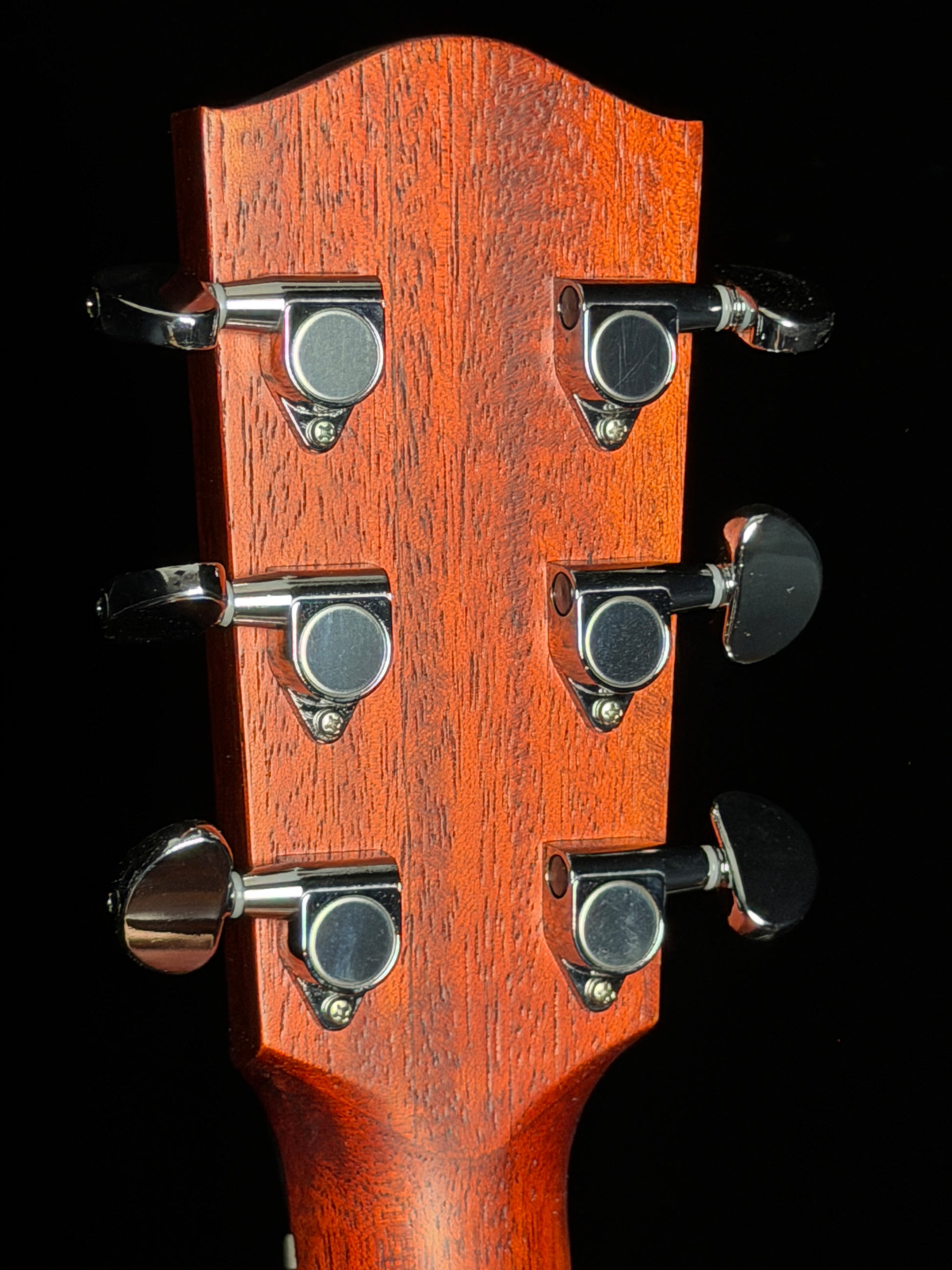 Close-up of a guitar headstock with six tuning pegs and a reddish-brown wood finish against a black background.