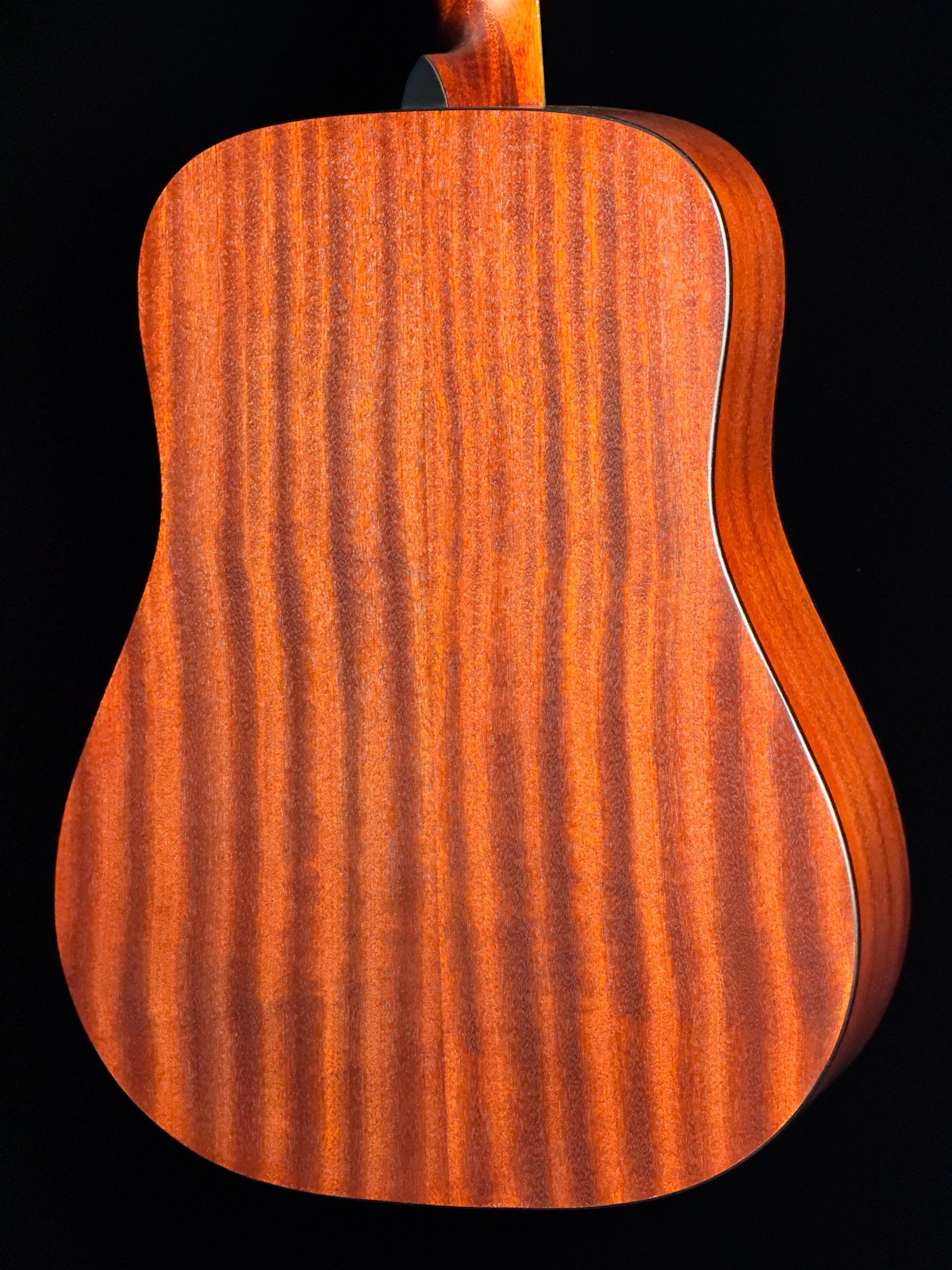 Close-up of the back of an acoustic guitar showing a rich, figured wood grain finish in warm reddish-brown tones against a solid black background.